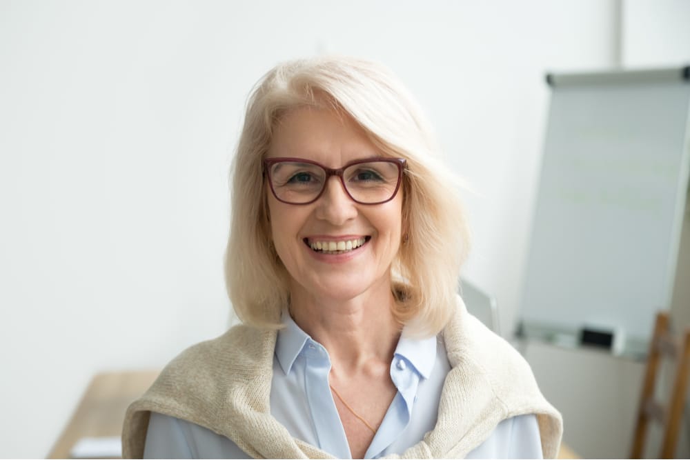 En leende äldre kvinna med axellångt vitt hår bär en ljusblå blus och glasögon. Hon har en lätt tröja draperad över axlarna. Hon står i ett modernt kontorsutrymme med en suddig whiteboardtavla och skrivbord i bakgrunden. Atmosfären framstår som ljus och välkomnande.